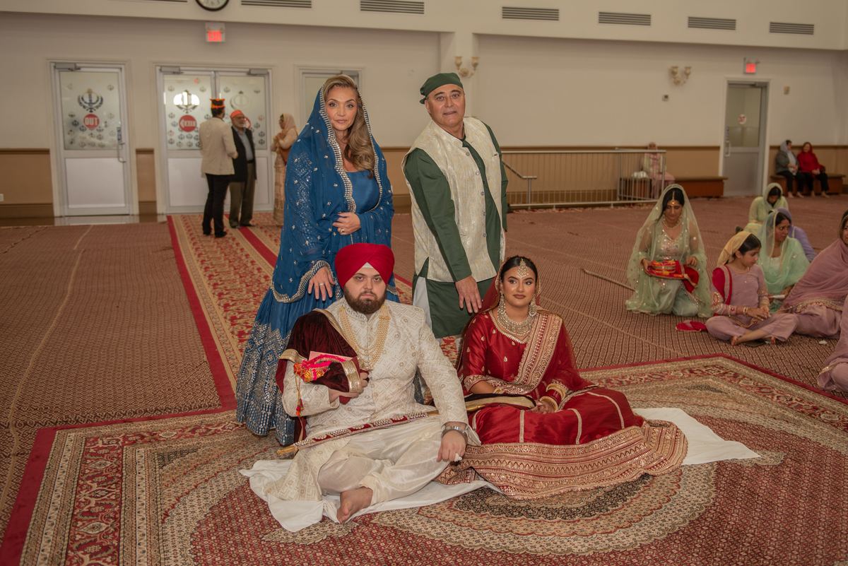 A Sikh wedding ceremony inside a gurdwara with the bride in red and groom in white, holding ceremonial items.