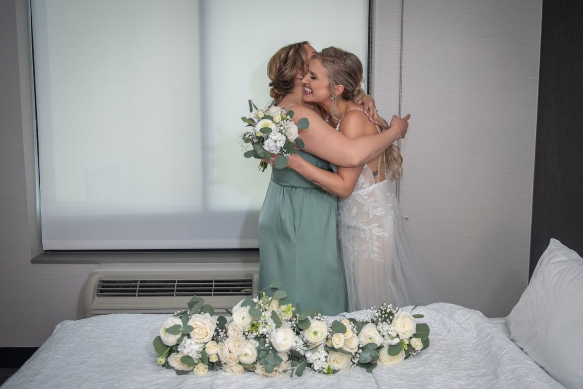 A bride in a white lace gown embraces a bridesmaid in sage green as they smile warmly near floral arrangements on the bed.
