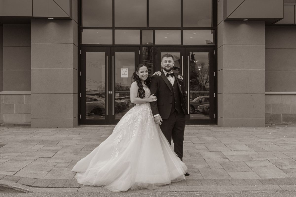 A smiling bride and groom pose outside a modern building with glass doors reflecting trees and cars in sepia tones.