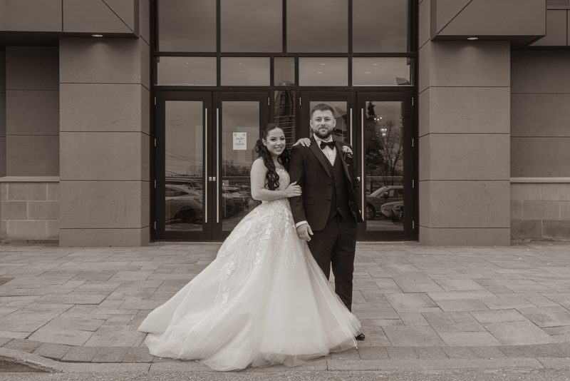 A smiling bride and groom pose outside a modern building with glass doors reflecting trees and cars in sepia tones.