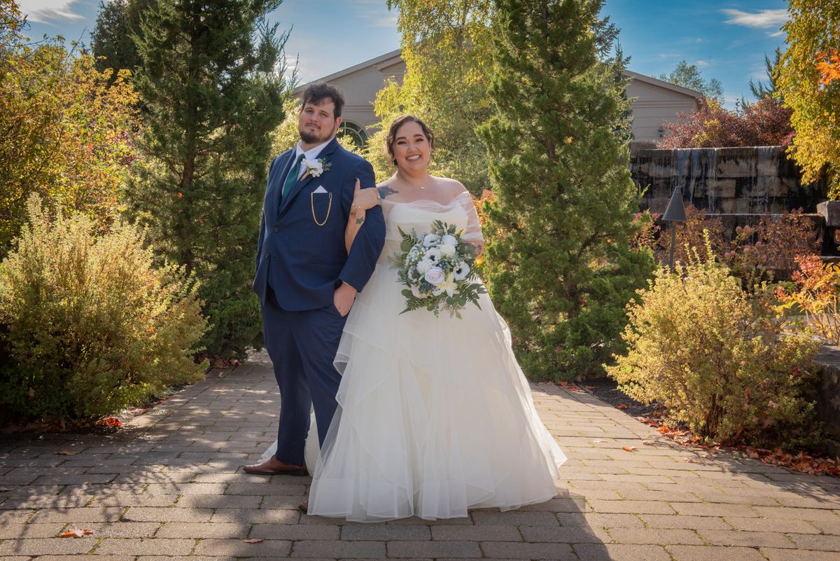A bride in a white gown and a groom in a navy suit stand smiling on a stone pathway surrounded by autumn trees.