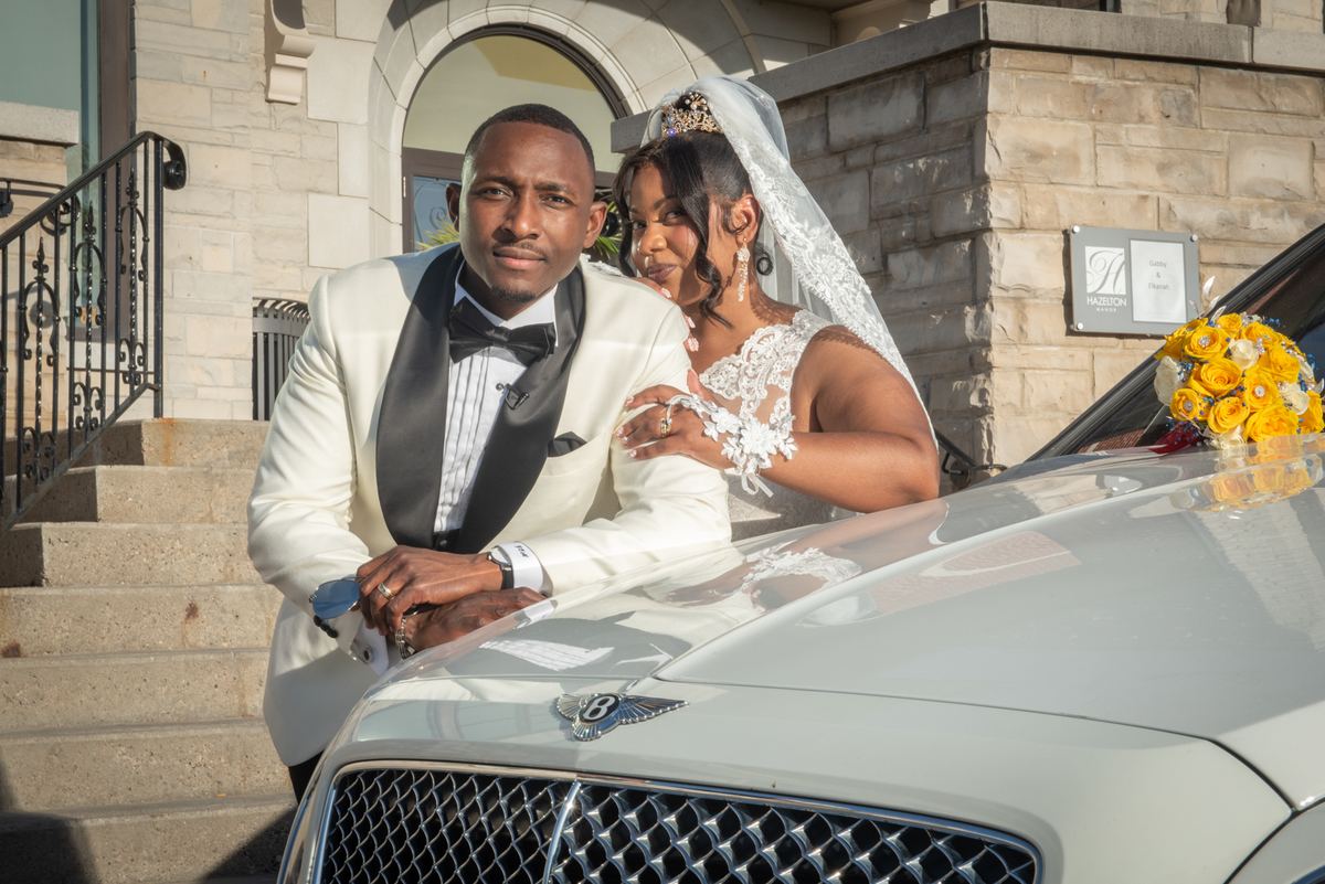 A newlywed couple in formal attire smiles beside a luxury car, with sunlight casting shadows on the grand building's steps.