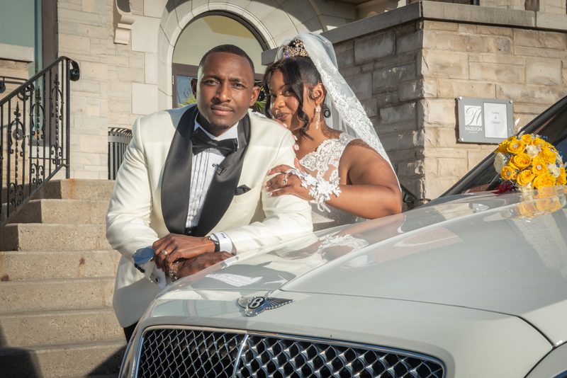 A newlywed couple in formal attire smiles beside a luxury car, with sunlight casting shadows on the grand building's steps.