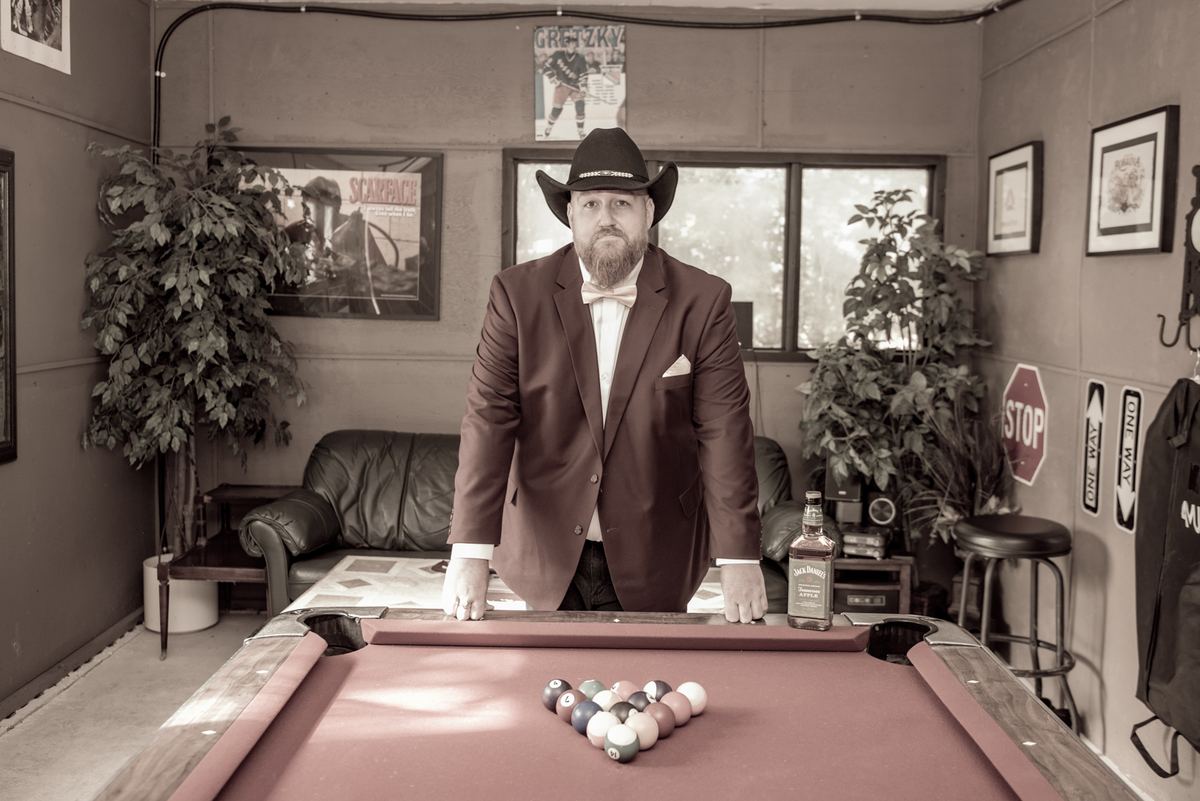 A man in a cowboy hat stands by a pool table in a sepia-toned room with posters and plants, awaiting play.