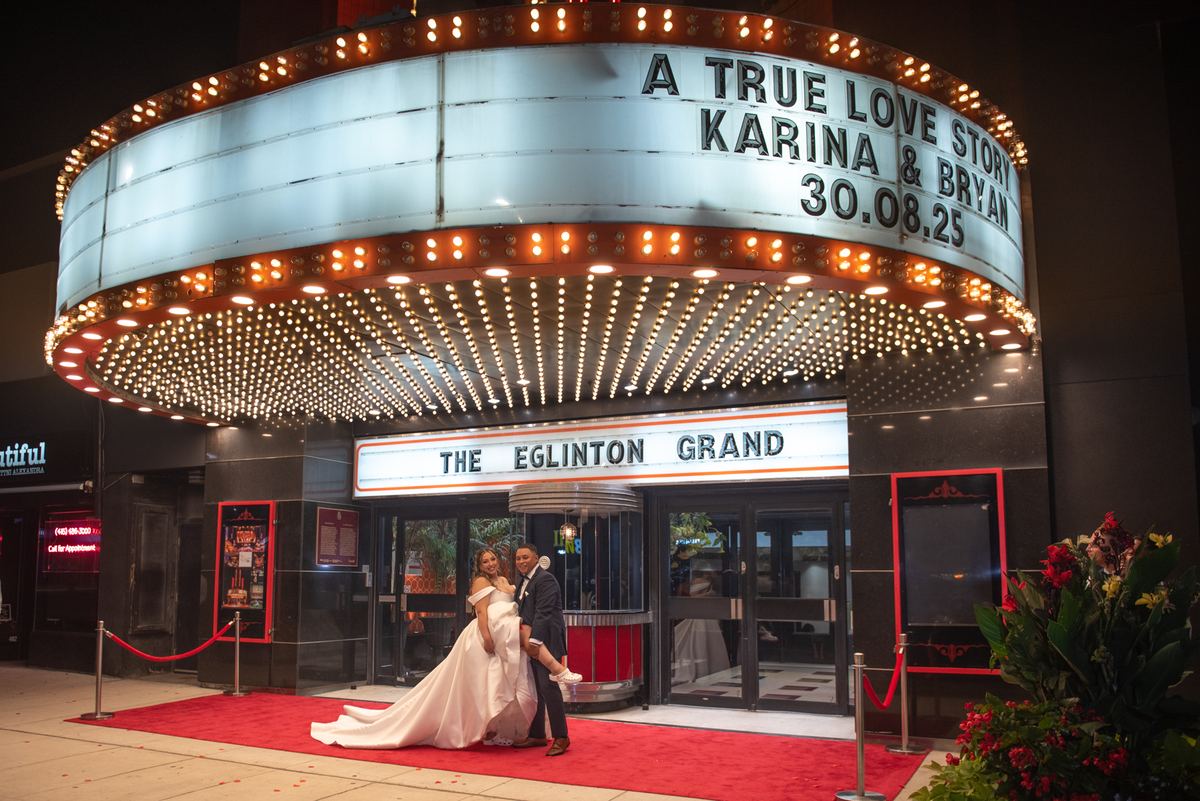 Karina and Bryan pose joyfully on a red carpet in front of a glowing marquee, celebrating their wedding.