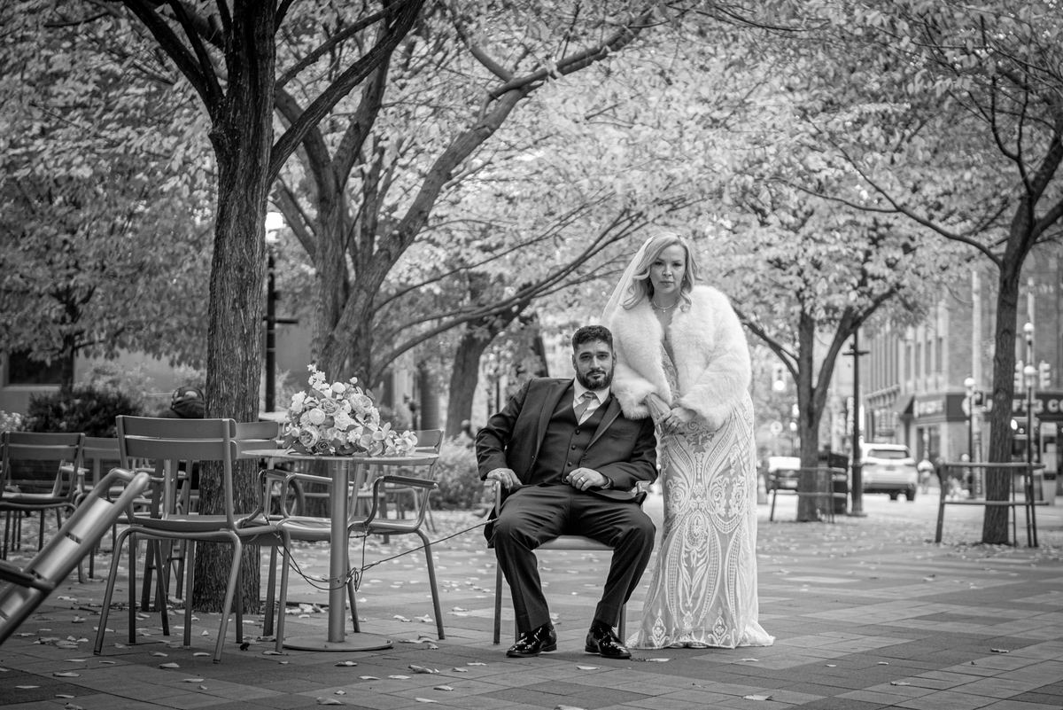 A newlywed couple poses in an urban plaza, the bride standing beside the seated groom with her hand on his shoulder.