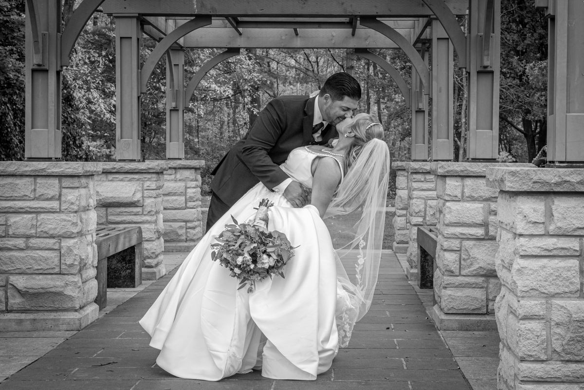 A bride and groom share an affectionate kiss under a wooden arbor, softly lit by natural daylight filtering through trees.