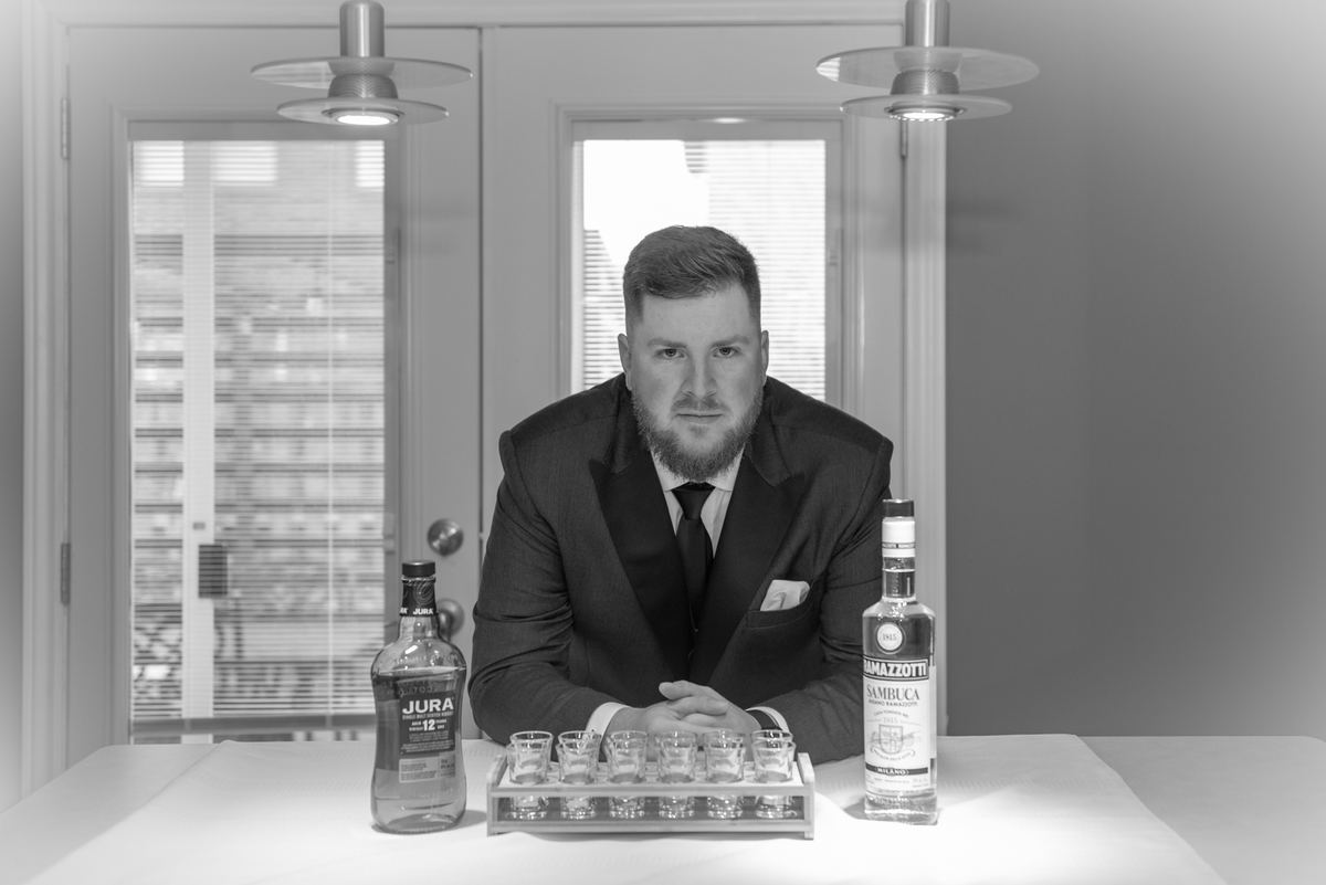 A man in a suit sits at a table with spirits and shot glasses, serious expression under soft lighting.