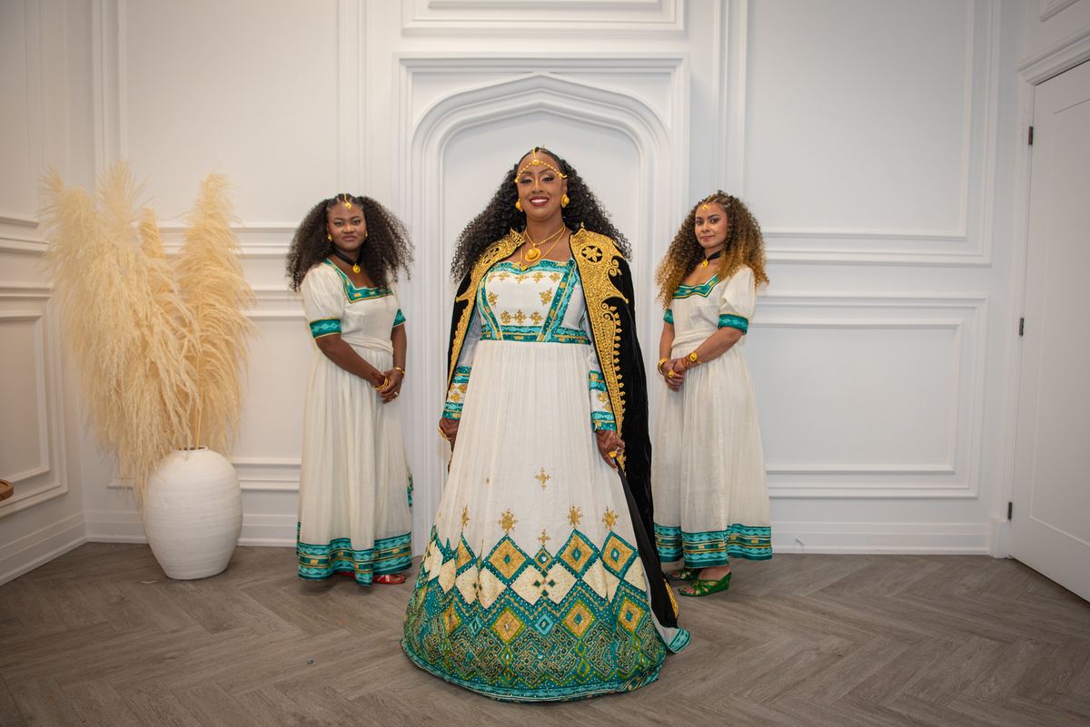 Three women in ornate dresses stand in a minimalist room, with the central figure in a white and teal gown smiling.
