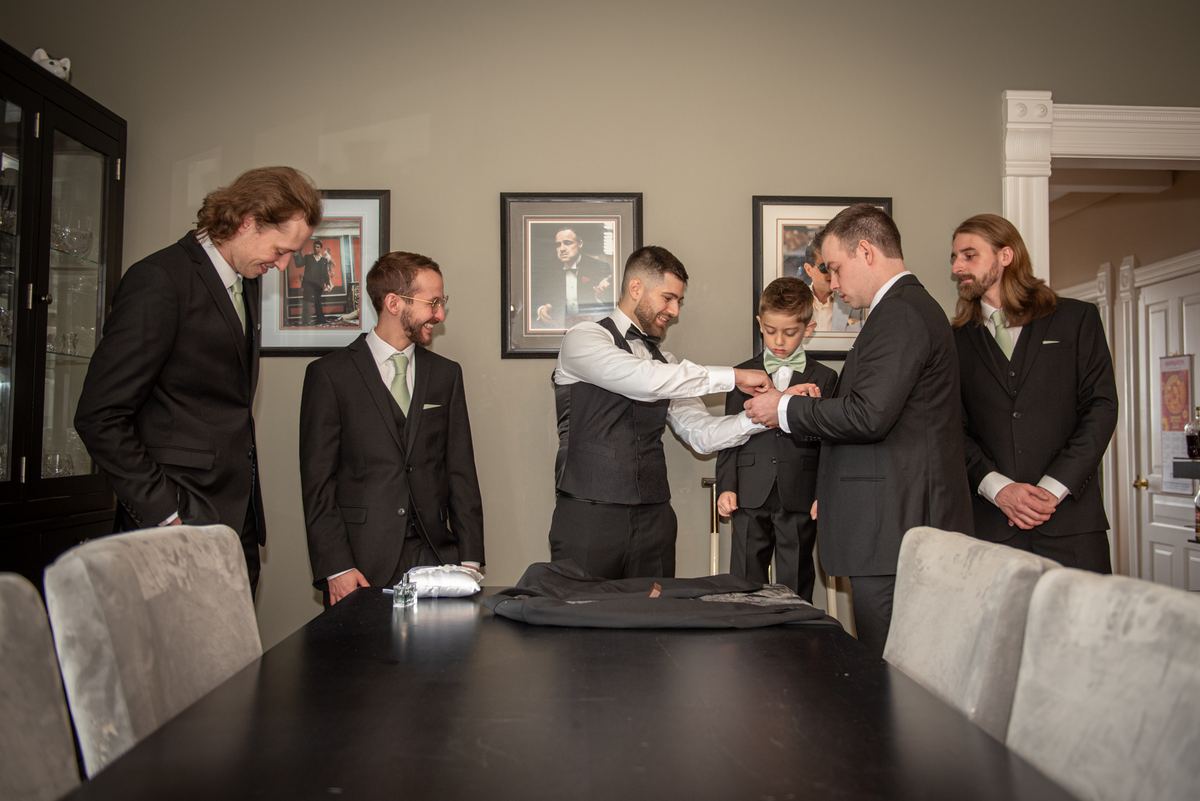 A groom and his party, including a young boy, gather around a table for pre-ceremony preparations in a warmly lit room.