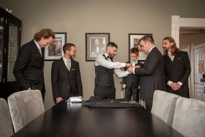 A groom and his party, including a young boy, gather around a table for pre-ceremony preparations in a warmly lit room.