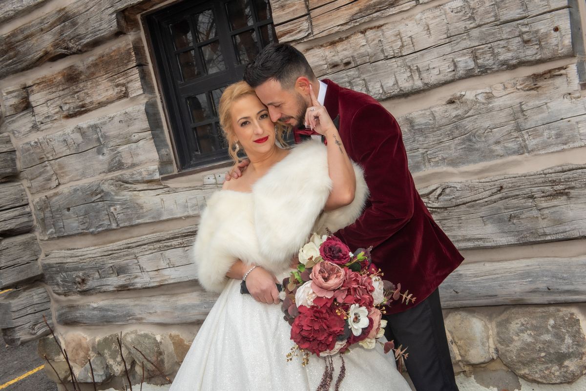 A bride in white and fur stole holds a bouquet as her groom in a burgundy jacket kisses her temple near a rustic log cabin.
