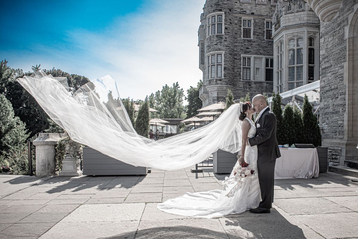A bride and groom kiss in a sunlit courtyard with a grand stone castle behind them, her veil billowing.