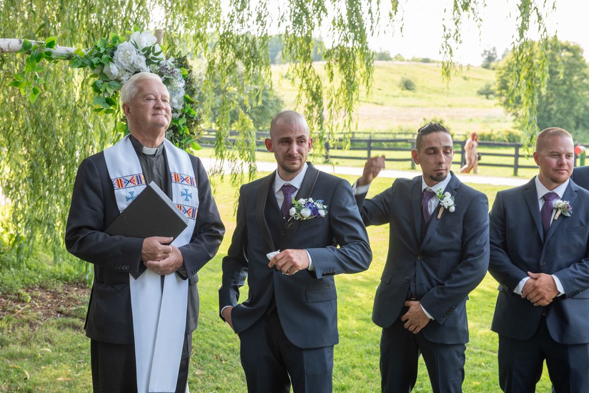 A wedding ceremony under a weeping willow arch covered in greenery and blooms, with the priest and groomsmen in dark suits.