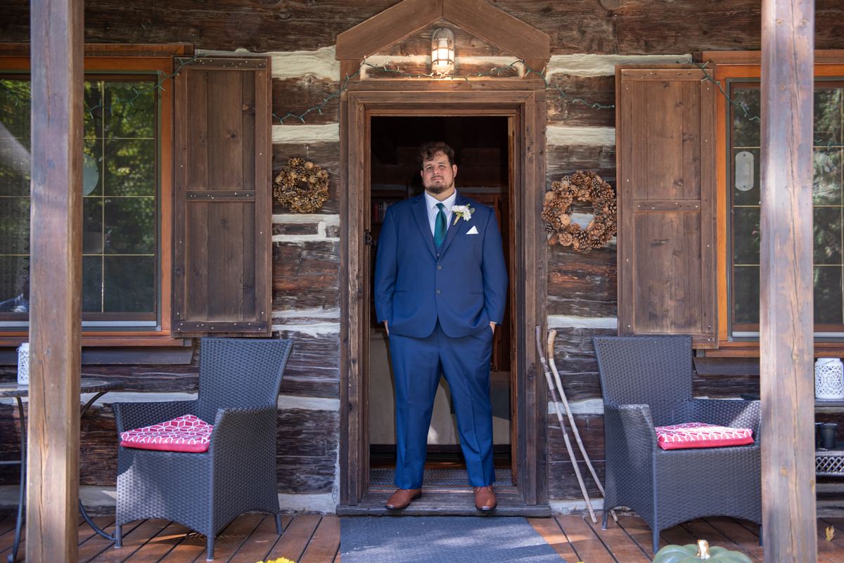 A groom in a blue suit stands at the cabin's porch door, looking at the camera, with warm daylight and autumn decorations.