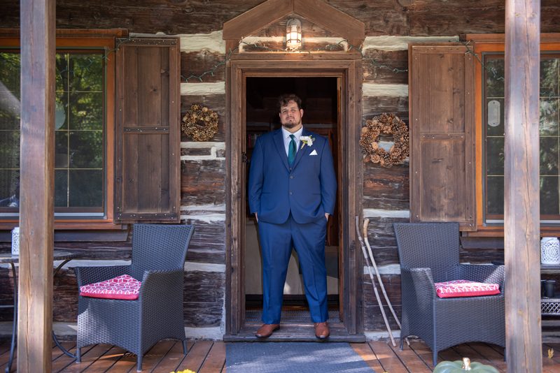 A groom in a blue suit stands at the cabin's porch door, looking at the camera, with warm daylight and autumn decorations.