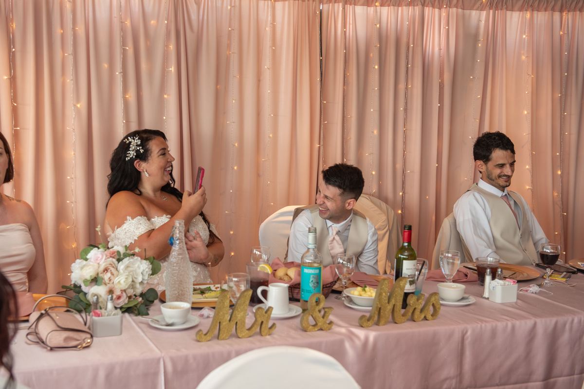 Bride laughs while holding her phone near groom at reception with string lights and floral arrangements.