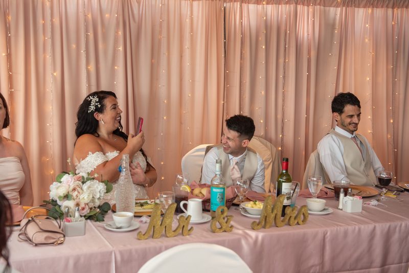 Bride laughs while holding her phone near groom at reception with string lights and floral arrangements.