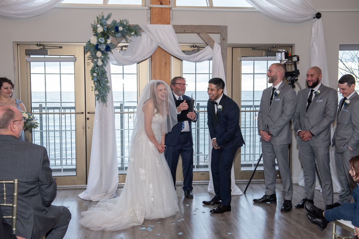 A bride in white laughs with her smiling groom as confetti falls on the wooden floor during their indoor ceremony.