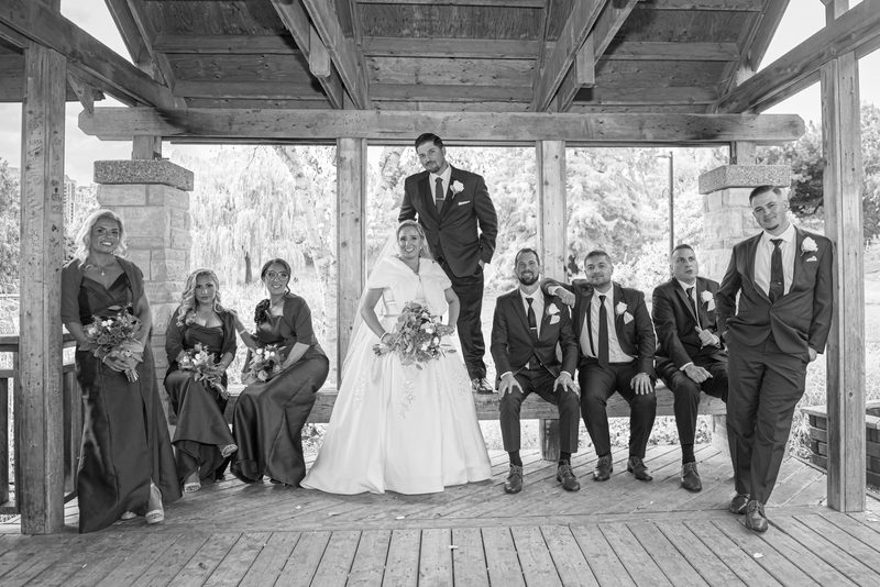 A wedding party poses together on a wooden deck under a rustic pergola, with the bride and groom at center.