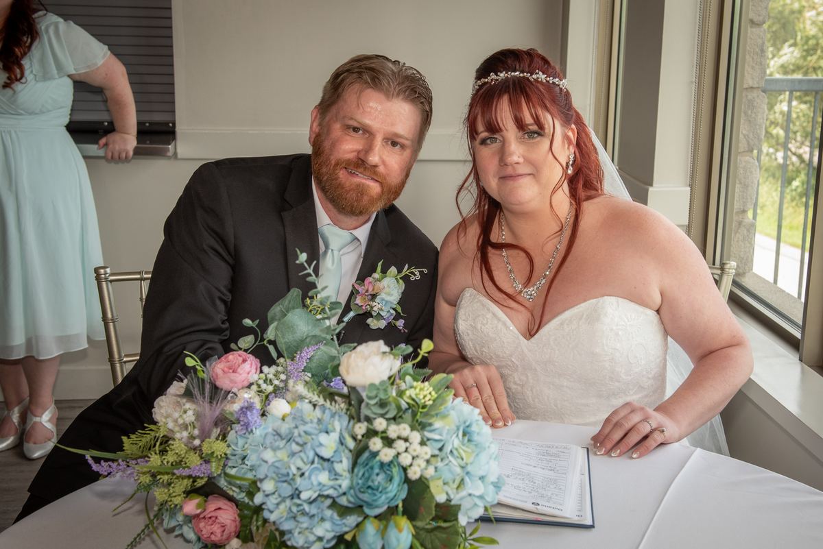 A newlywed couple smiles as they sign documents at a table with a floral arrangement, illuminated by soft natural light.