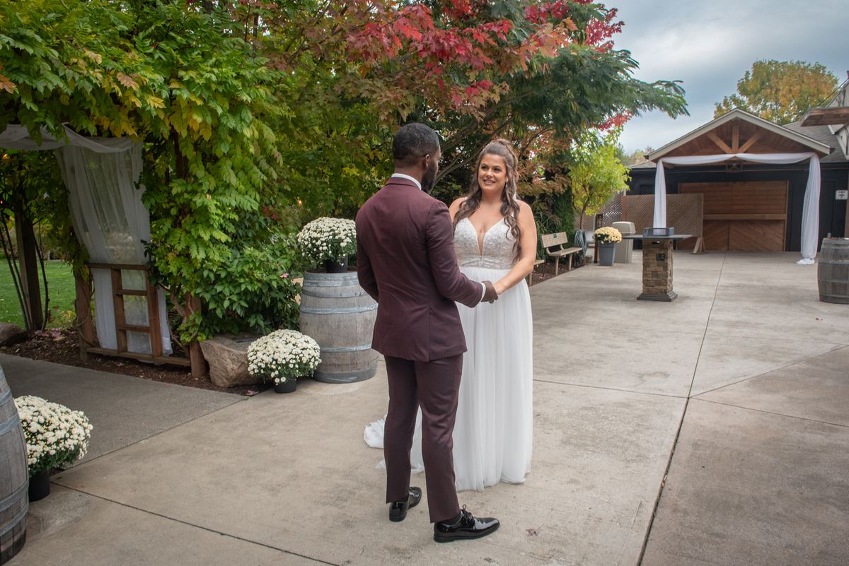 A bride and groom stand hand-in-hand in an autumn courtyard, smiling tenderly as rustic decor frames them.