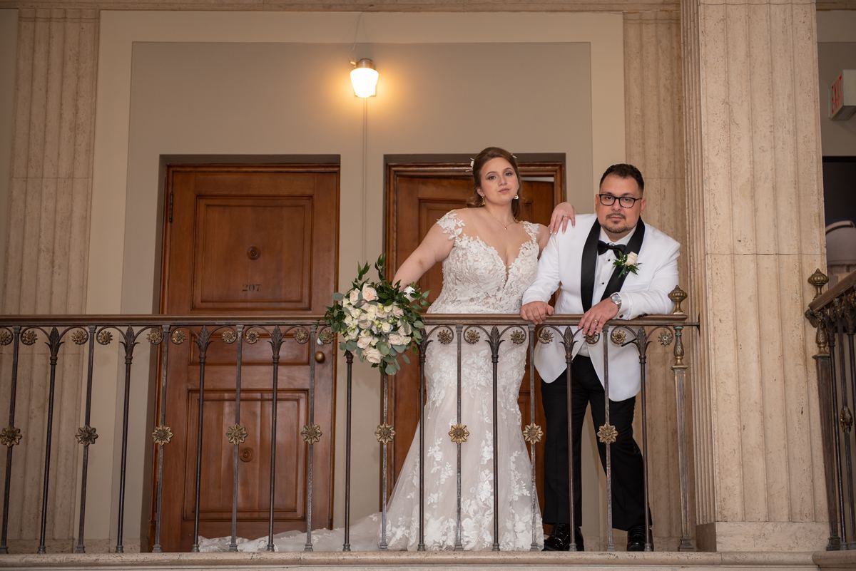 A bride and groom pose on an ornate balcony, illuminated by a pendant light, under the warm glow of their wedding day.