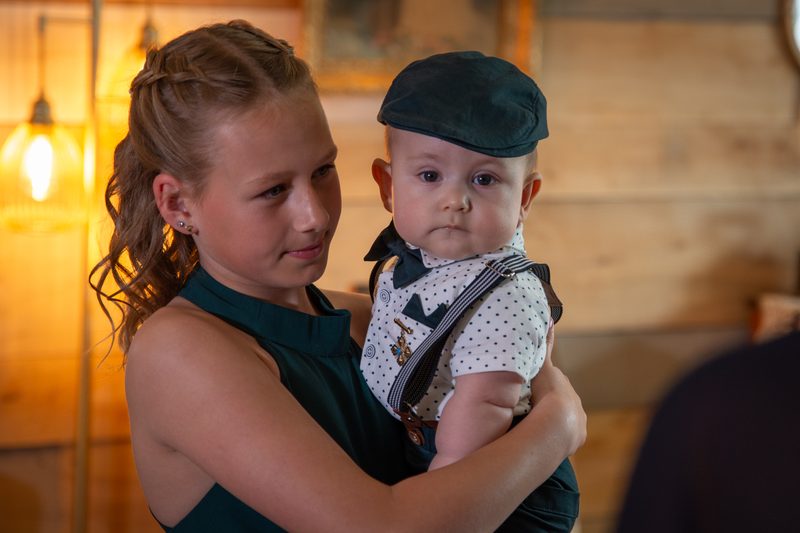 A young girl holds a formally dressed baby with tender expressions in warm, golden light against wooden panels.