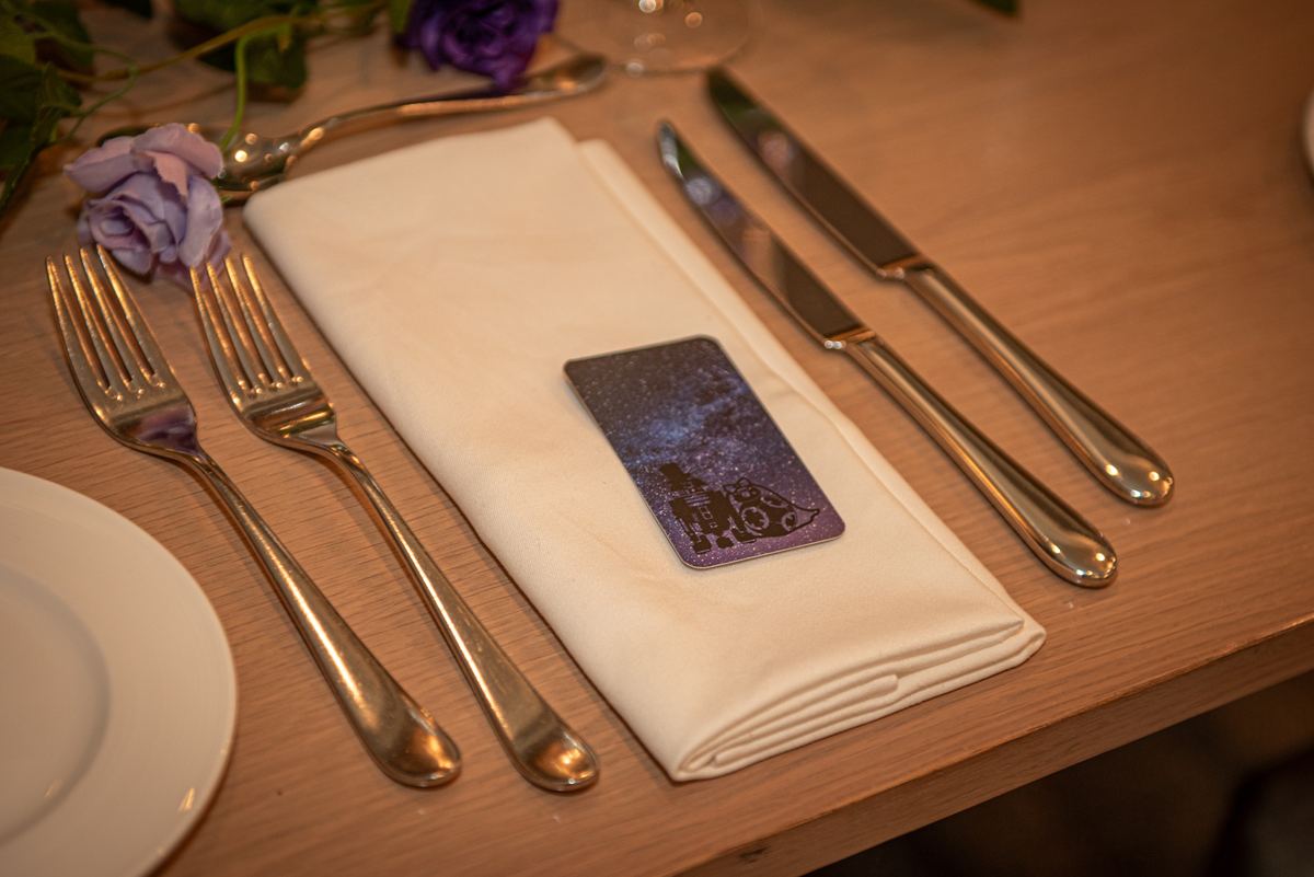 Elegant table setting with silverware, folded napkin, decorative card, and purple roses under warm lighting.