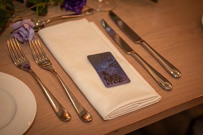 Elegant table setting with silverware, folded napkin, decorative card, and purple roses under warm lighting.