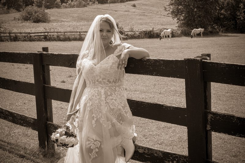 A bride in a lace gown leans against a wooden fence, smiling with her bouquet as horses graze in the background.