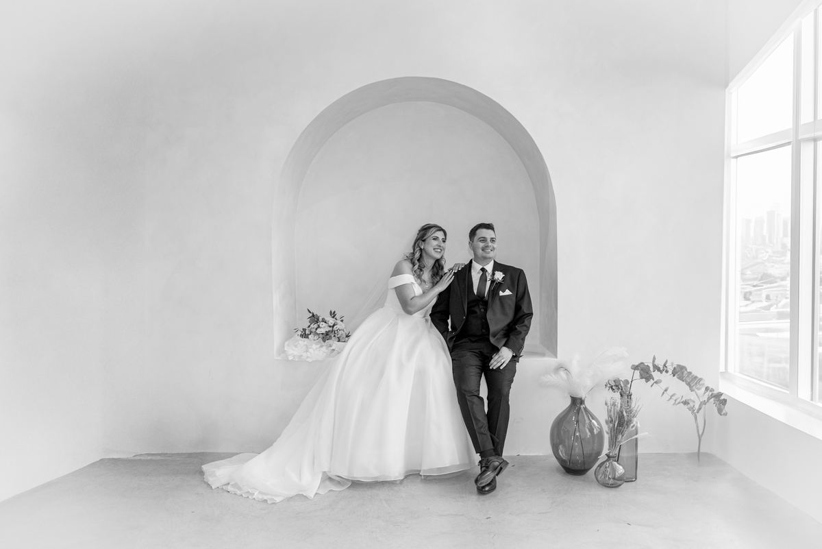 A smiling bride and groom sit beside an arched alcove in a bright white room, illuminated by natural light.