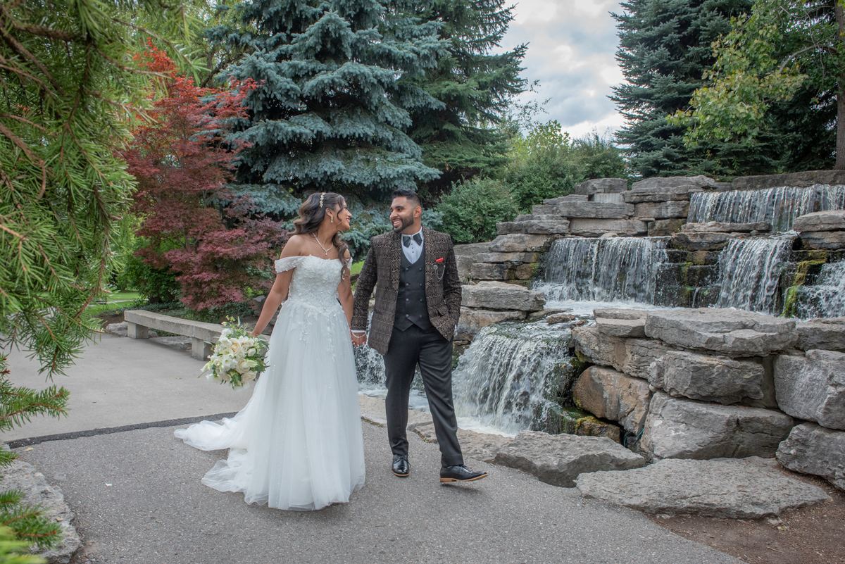 A newlywed couple walks hand-in-hand beside a stone waterfall, smiling at each other under an overcast sky.