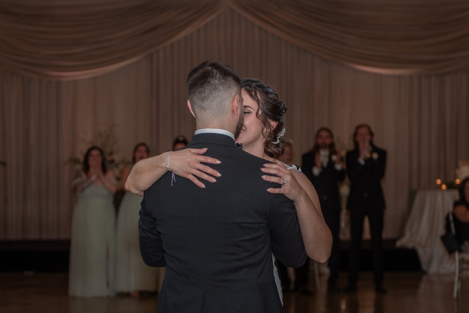 Seen from behind, a bride and groom sway in their first dance under warm light while guests clap and film from the tables.