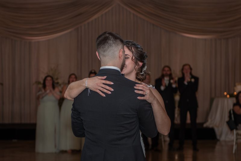 Seen from behind, a bride and groom sway in their first dance under warm light while guests clap and film from the tables.