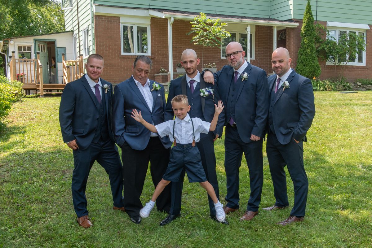 A young boy poses energetically between five groomsmen in dark suits, all smiling on a sunlit lawn in front of a house.