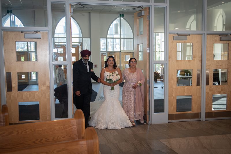 A bride in a white lace gown walks through wooden doors into a modern chapel, escorted by family members.