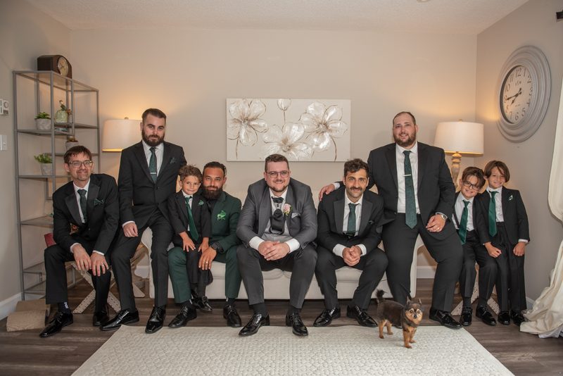 A groom and his groomsmen, along with young boys, pose in matching dark suits and green ties in a warmly lit living room.