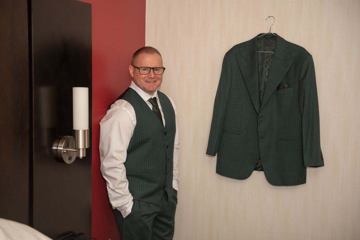 A man in a pinstripe suit and vest leans against a wall with a jacket hanging nearby, suggesting readiness for an event.