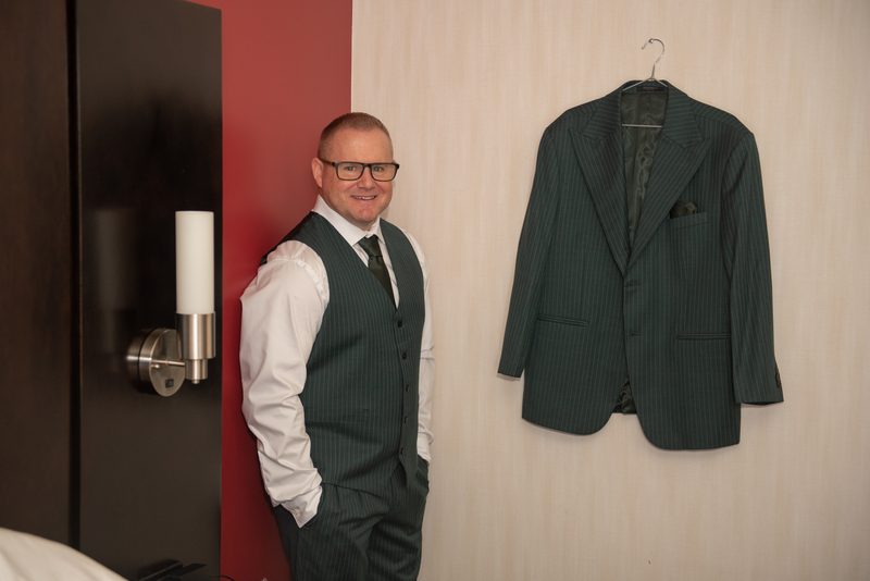 A man in a pinstripe suit and vest leans against a wall with a jacket hanging nearby, suggesting readiness for an event.