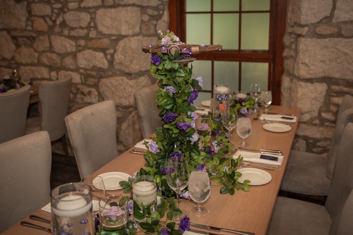 A rustic dining table set with greenery, purple flowers, and floating candles in soft light from a wooden window frame.