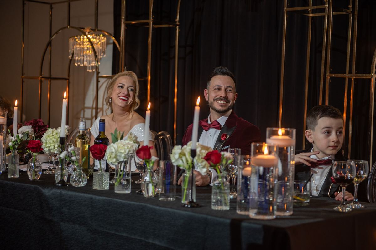 A bride and groom smile from the head table beside a young boy in a tuxedo, lit by candlelight and a chandelier overhead.