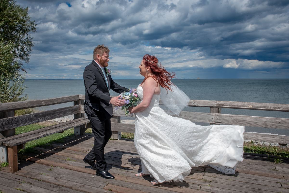 A bride and groom walk hand in hand on a wooden boardwalk, the bride's dress billowing in the wind as stormy clouds loom.
