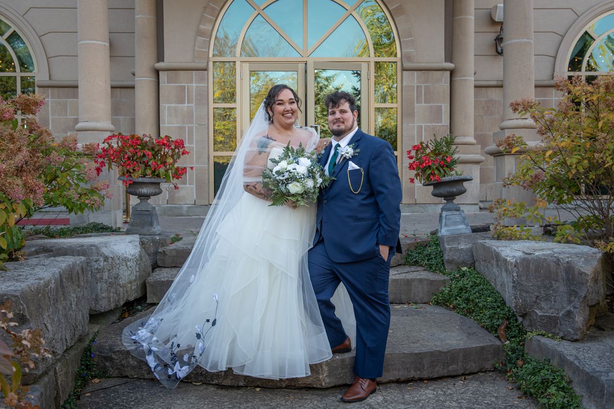 A bride and groom stand on stone steps outside an elegant building, surrounded by autumn foliage and red flowers.