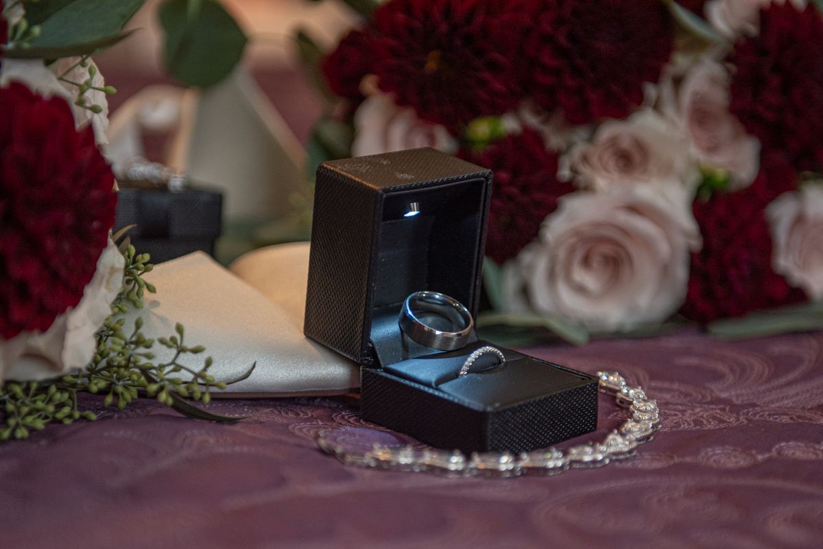 Wedding rings in an open black box beside a sparkling bracelet on burgundy fabric with blurred flowers in the background.