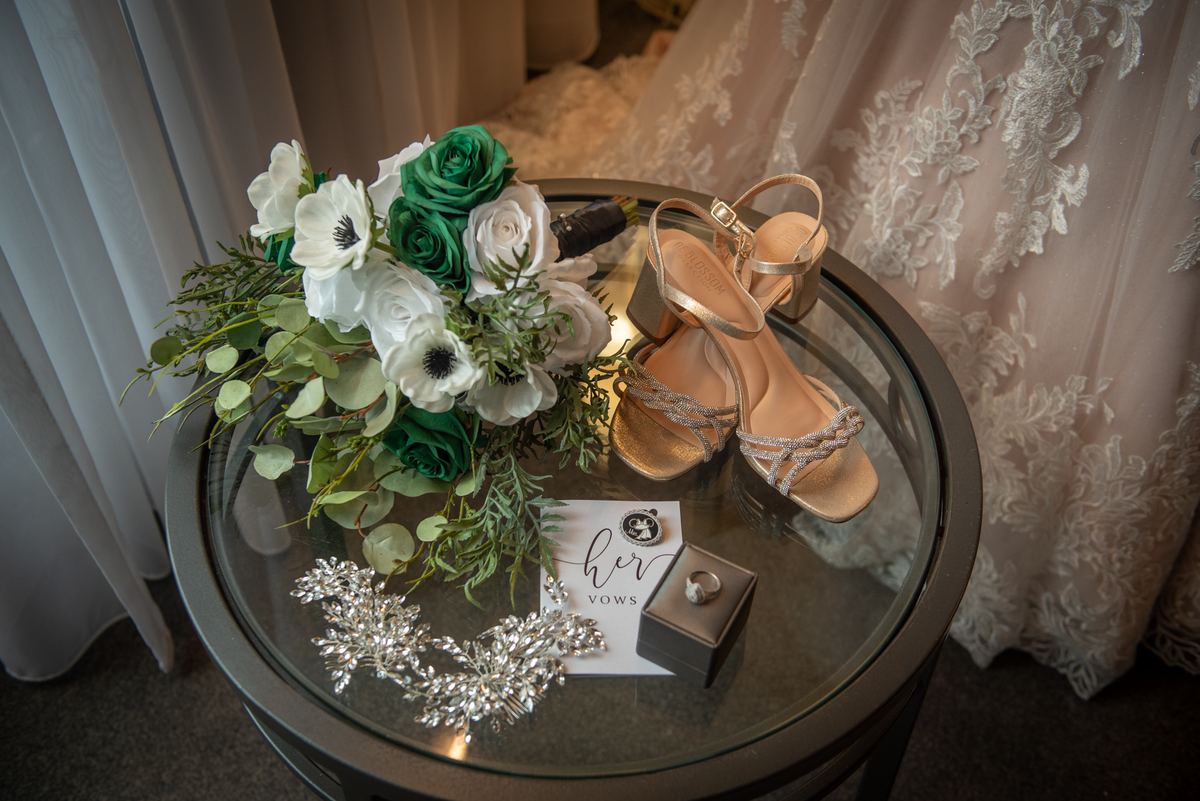 A glass table holds a wedding bouquet, heels, ring box, and hairpiece beside a lace gown under soft light.