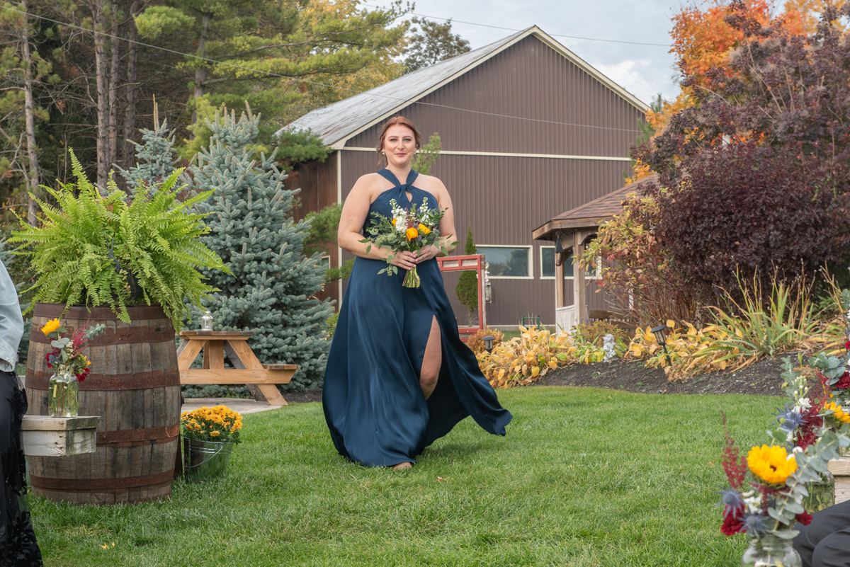 A woman in a navy-blue dress walks across a grassy lawn toward the camera, holding a bouquet, with autumn foliage.