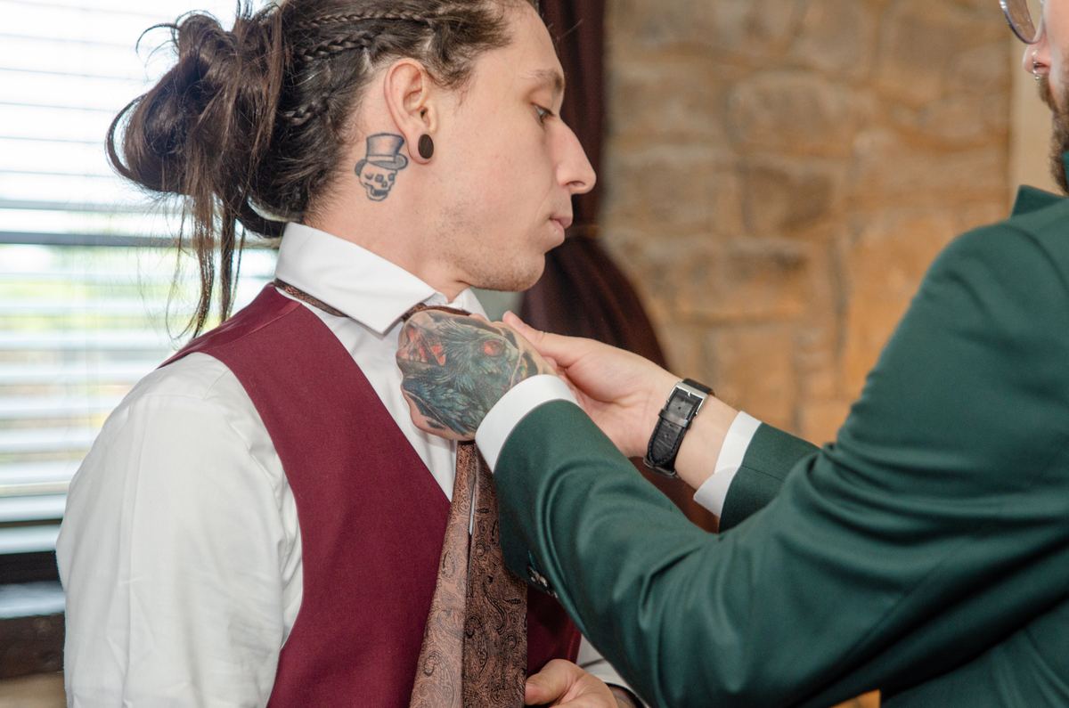 A groom with a skull tattoo and braided hair is adjusting his paisley tie with help from another man.