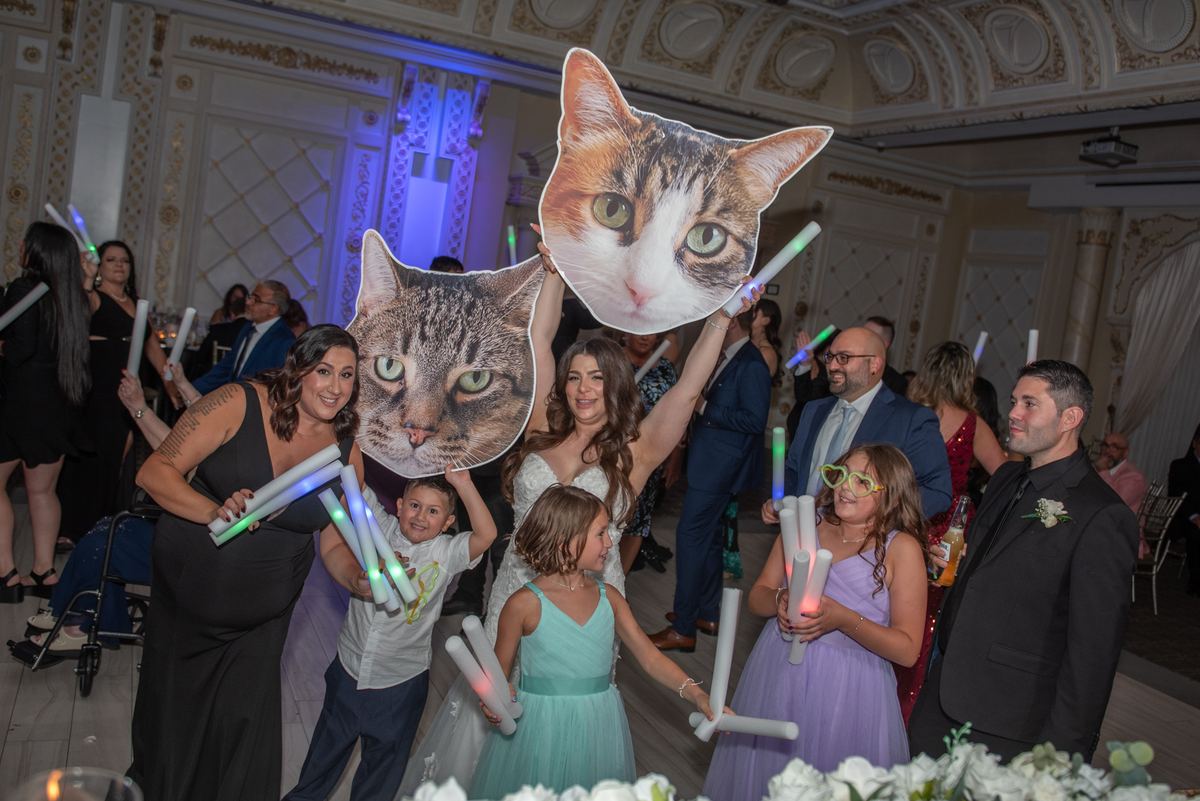 Guests, including the bride and children, pose joyfully with cat-face cutouts and light sticks at a wedding reception.