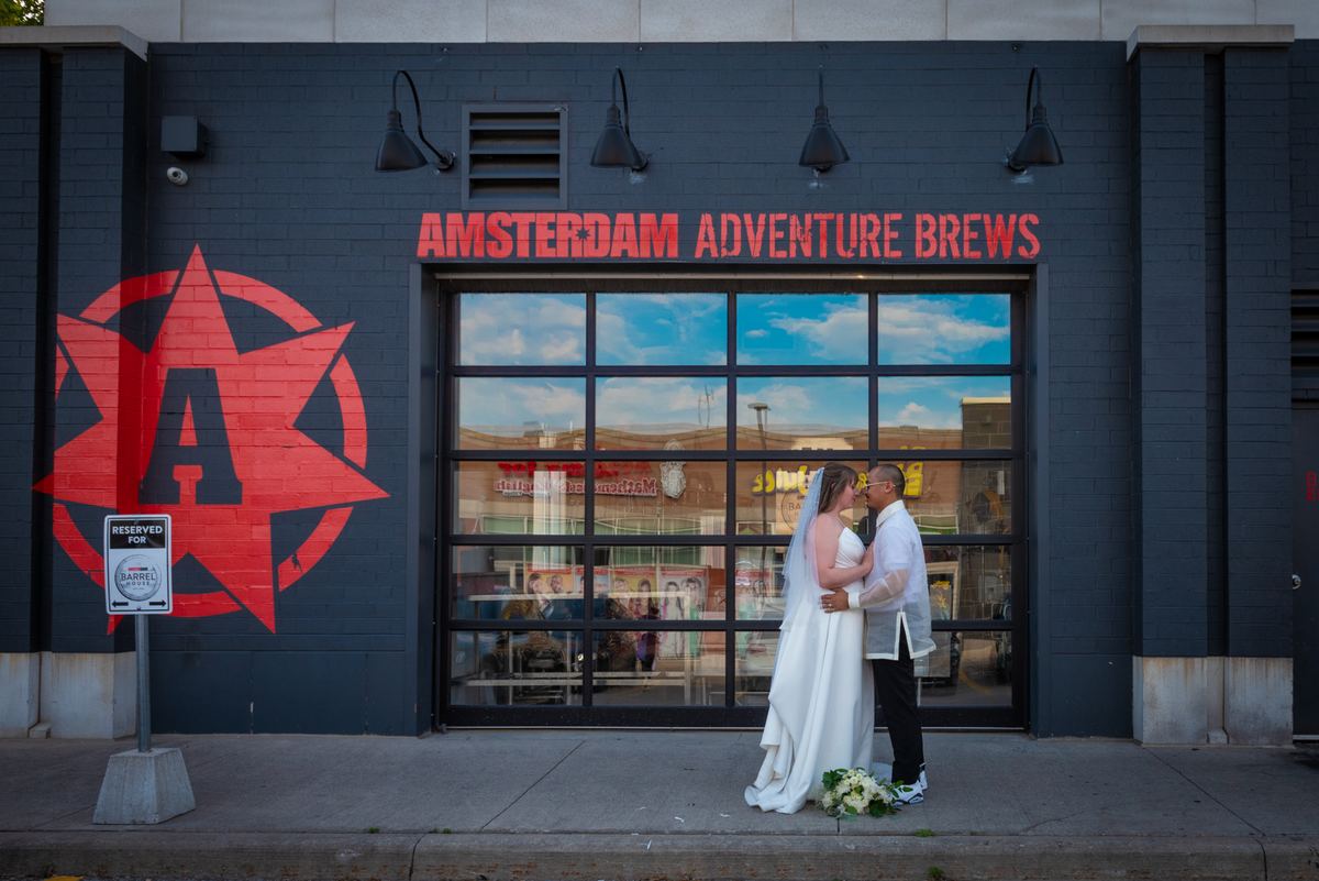 A bride and groom kiss on a sidewalk in front of a brewery with red branding, under golden late-afternoon light.
